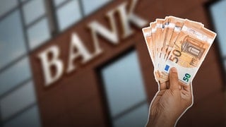 Bankgebäude im Hintergrund und eine Hand hält 50 Euro Scheine. Zinsvergleich – darauf sollten Sparer jetzt achten