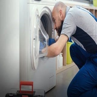 Handwerker mit Glatze und Bard trägt einen Blaumann und sitzt auf dem Boden rechts im Bild. Er repariert eine Waschmaschine (links im Bild).