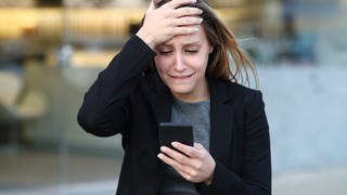 Eine Frau guckt aufgebracht auf ihr Handy, da sie Opfer von Sim Swapping geworden ist. Die Betrugsmasche kann nervenaufreibend und teuer werden! Wie Betrüger Handynummern stehlen und wie ihr euch gegen SIM Swapping schützen könnt. 
