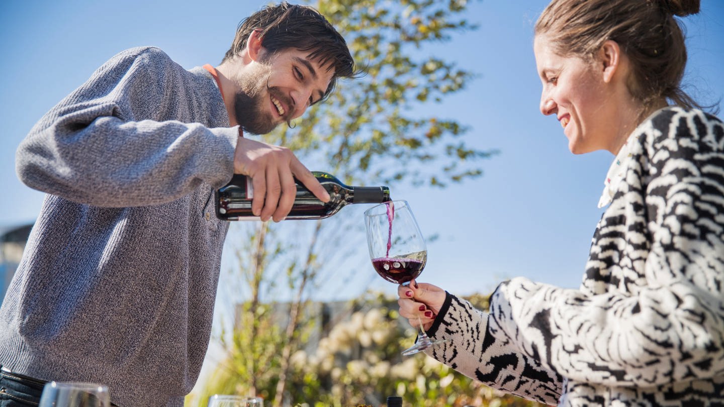 Zwei Menschen im Bild schenken sich Wein ein. Picknick im Freien. Freizeit an Pfingsten schön gestalten.