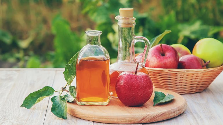 Auf einem Tisch steht ein Glas mit Apfelessig und ein Apfel daneben. Abnehmen, schöne Haut, tolle Haare – wie sinnvoll ist Apfelessig für die Gesundheit? Wie gut sind die Inhaltsstoffe und was ist mit anderem Essig? 