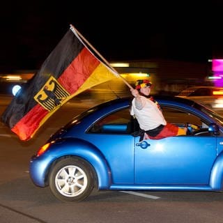Junger Mann mit riesiger Deutschlandflagge sitzt bei voller Fahrt auf Autofenster von blauem Kleinwagen. Was ist beim Autokorso erlaubt? Darf ich Zimmer an Fußballfans untervermieten?