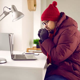 Ein Mann trägt eine rote Wollmütze auf seinem Kopf. Außerdem trägt er Handschuhe und eine Winterjacke. Er sitzt auf einem Stuhl, vor einem Schreibtisch, auf dem ein Laptop steht. Seine Hände hält er sich zum wärem vor den Mund.