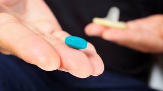 Auf einer Hand liegt eine blaue Viagra Pille, die gegen Erektionsprobleme helfen soll. Im HIntergrund ist ein Kondom zu sehen.
