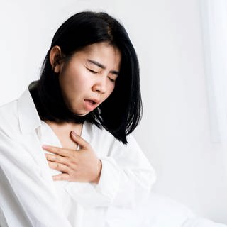 Asiatische Frau mit Panikstörung im Bett, die sich mit der Hand die Brust hält und kaum atmen kann