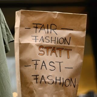 Demonstranten halten in Stuttgart ein T-Shirt mit der Aufschrift "Mir ist nicht egal wer dieses T-Shirt gemacht hat" und eine Tüte mit der Aufschrift "Fair Fashion statt Fast Fashion" hoch.