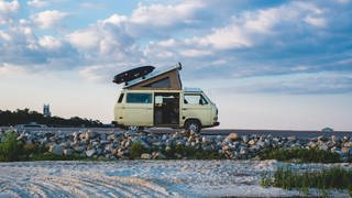 Beiger VW Bully steht hinter einem Steinstrand mit aufgestelltem Aufstelldach und montiertem Jetbag. Wie ist man auf Reisen mit Wohnmobil und Camper besonders nachhaltig und umweltfreundlich unterwegs?