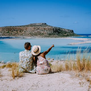 Pärchen in Strandkleidung sitzt auf sandiger Anhöhe und blickt auf die Lagune von Balos auf Kreta. Was kostet Urlaub auf der griechischen Insel?