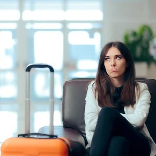 Eine Frau sitzt beleidigt mit ihrem Koffer am Flughafen. Gepäck verloren auf der Reise oder schlechtes Hotel: Ihre Rechte im Urlaub bei Reisemängeln und welche Entschädigung Ihnen zusteht.