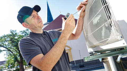 Ein Handwerker installiert eine Klimaanlage an einer Hauswand. Wieder eine Hitzewelle. Doch das Kühlen per Gerät kann teuer werden und dem Klima schaden. Monoblock, Luftkühler oder Ventilator: Was ist sinnvoll und wie geht es ohne Strom? 