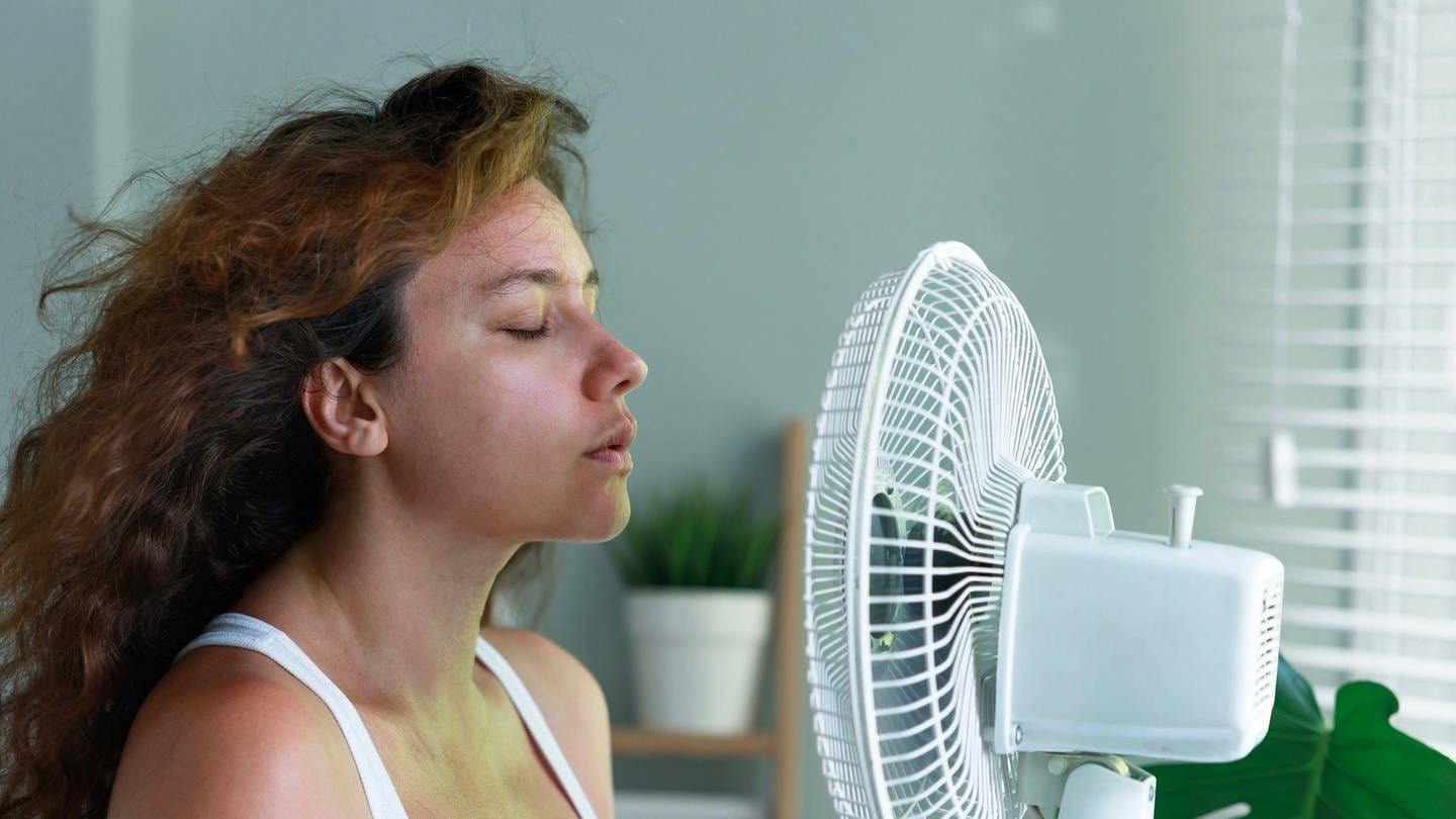 Eine Frau sitzt vor einem Ventilator und genießt den Luftzug. Wieder Hitzewarnung: Doch eine Klimaanlage für eine kühle Wohnung kann teuer werden und dem Klima schaden. Was ist sinnvoll und wie geht Kühlen auch ohne Klimaanlage?