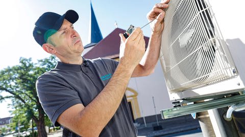 Ein Handwerker installiert eine Klimaanlage an einer Hauswand. Wieder eine Hitzewelle. Doch das Kühlen per Gerät kann teuer werden und dem Klima schaden. Monoblock, Luftkühler oder Ventilator: Was ist sinnvoll und wie geht es ohne Strom?