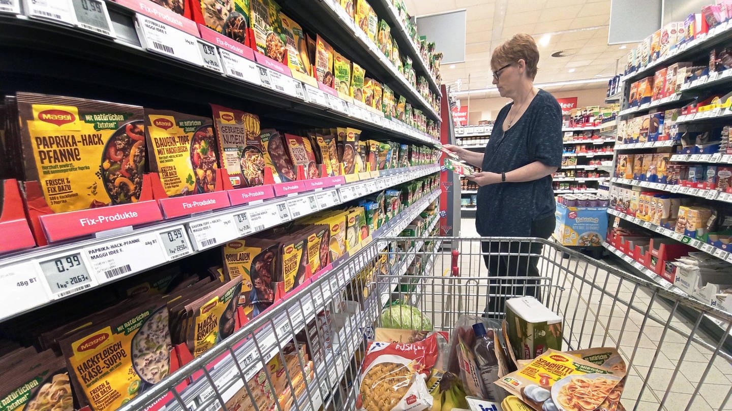 Frau mit Einkaufswagen im Supermarkt vor dem Regal mit Produkten von Maggi: Maggi Würze steht seltener auf dem Tisch, aber meterweise Maggi-Produkte im Supermarkt - trotz schlechtem Ruf von Fertiggerichten. Vier Wochen nur Maggi essen: Was passiert dann?