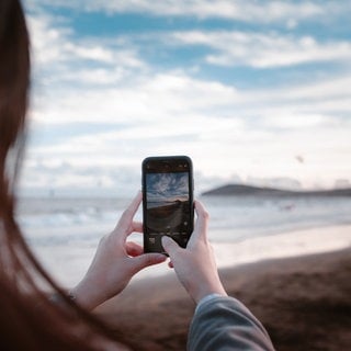 Frau fotografiert mit dem Smartphone das Meer. Bessere Bilder mit dem Handy machen: Wir zeigen Hacks, Tricks und Tipps.