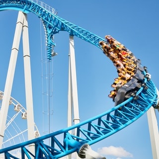 Eine blaue Achterbahn hebt sich vom ebenfalls blauen Himmel ab. Ein Wagon rast um eine Kurve.