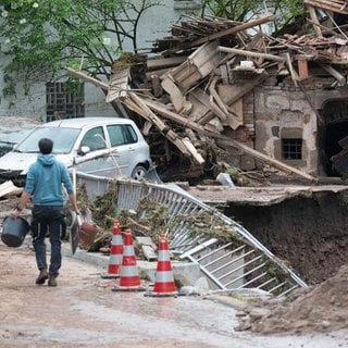 Ein von Hochwasser stark zerstörtes Gebäude neben Erdrutsch, davor ein Mann mit Eimer.