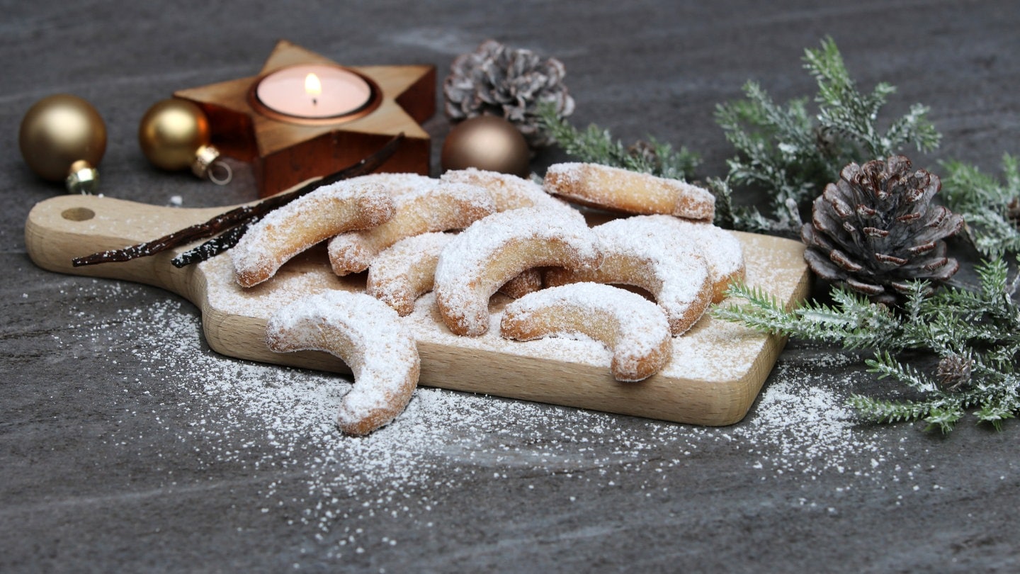 Vanillekipferl liegen neben Tannenzweigen und Kerze auf einem Holzbrett. Wie gelingen Weihnachts-Plätzchen möglichst gesund? wie lässt sich teure Butter ersetzen?