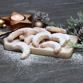 Vanillekipferl liegen neben Tannenzweigen und Kerze auf einem Holzbrett. Wie gelingen Weihnachts-Plätzchen möglichst gesund? wie lässt sich teure Butter ersetzen?