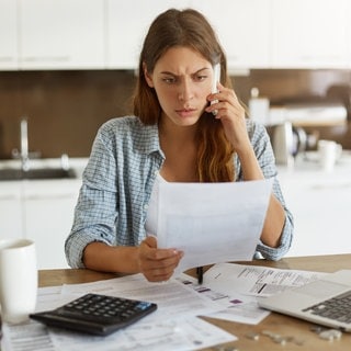 Eine besorgte junge Frau sitzt an einem Schreibtisch voller Rechnungen und telefoniert. Energetische Sanierung, Modernisierung – klingt gut: Die Wohnung soll energiesparender und schöner werden. Für Mieter heißt das oft: Mieterhöhung. Was dürfen Vermieter umlegen? Gibt es Obergrenzen?