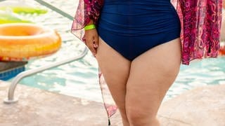 Eine korpulente Frau mit blauem Badeanzug posiert am Strand. Überproportional dicke Beine im Vergleich zum restlichen Körper können durch ein Lipödem bedingt sein.