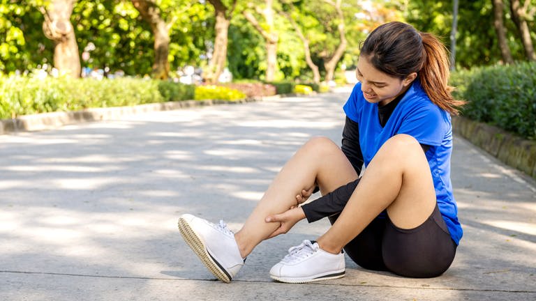 Eine Sportlerin in Joggingschuhen sitzt mit Schmerzen in der Wade auf dem Boden - Muskelkrämpfe, viele Sportler gingen bisher davon aus, dass ein Mangel an Magnesium die Ursache ist.