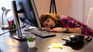Junge Frau schläft vor ihrem Computer mit dem Kopf auf dem Tisch. Was bringen Mittagsschlaf und Powernap gegen Müdigkeit?