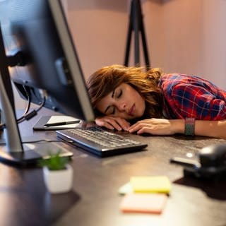 Junge Frau schläft vor ihrem Computer mit dem Kopf auf dem Tisch. Was bringen Mittagsschlaf und Powernap gegen Müdigkeit?