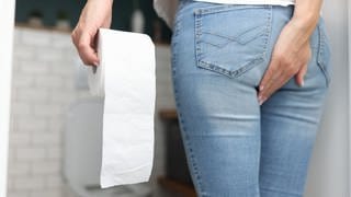 Frau steht in einem Badezimmer, trägt eine Jeans und und ein weißes Oberteil. In ihrer linken Hand hält sie eine Klorolle, mit der Rechten fasst sich sich an das Hinterteil. Im Hintergrund ist die Toilettenschüssel.