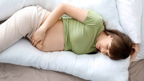 Eine schwangere Frau schläft auf der Seite. Ein U-förmiges Kissen stützt ihren Kopf und ihren Rücken, sodass sie nicht in die gefährliche Rückenlage kommt.