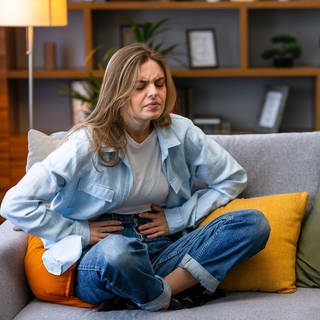 Eine Frau sitzt mit Bauchschmerzen auf einem Sofa und hält ihren Bauch mit den Händen.