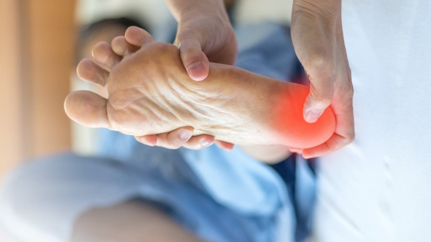 An einem Fuß ist die Fußsohle an der Ferse rot markiert, dort wo der Patient Schmerzen hat. Viele leiden an einer entzündeten Fußsohlensehne und Fersensporn. Wieder schmerzfrei laufen: einfache Übungen und Therapien ohne Operation.