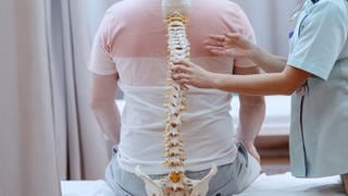 Gezeigt wird ein Patient, der mit dem Rücken zum Betrachtenden auf einer Liege sitzt. Eine Ärztin hält ein Skelettmodell einer Wirbelsäule an seinen Rücken.