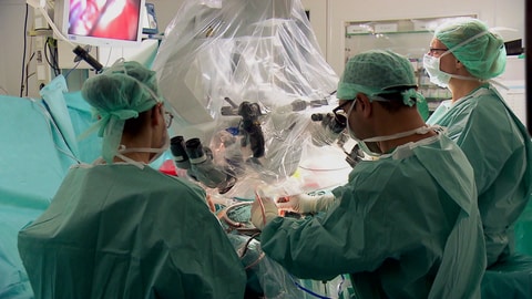 Eine Person wird in einem OP-Saal am Gehirn operiert, um sie herum stehen zwei Ärzte.