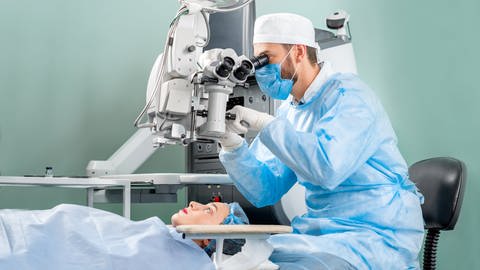 Ein Augenarzt in Operationskleidung sitzt an einem großen Mikroskop und schaut sich die Augen einer Patientin an. Er bereitet sich auf eine Laser-Operation der Augen  vor.