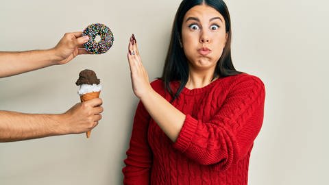 Frau mit dunklen Haaren und rotem Pullover lehnt einen Donut und ein Eis ab. Hunger, Sättigung, Appetit: Manchmal essen wir mehr als nötig und fühlen uns dennoch nicht satt. Neben der Ernährung sind soziale und psychologische Faktoren wichtig für den Appetit.