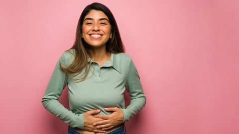 Eine Frau mit dunkelblonden langen Haare, Jeans und einer grünen Bluse greift sich an den Bauch und fühlt sich satt. Hunger, Appetit und Sättigung werden teils auch durch soziale und psychologische Faktoren bestimmt.