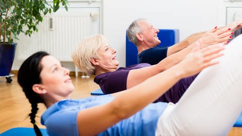 Eine ältere Frau und ein älterer Herr machen zusammen mit einer Trainerin eine Pilatesübung in Rückenlage.