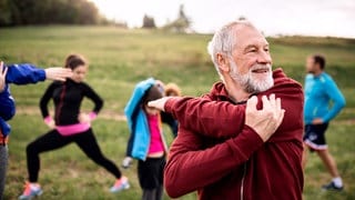 Ein älterer Mann macht Fitnessübungen, im Hintergrund ist eine Gruppe weiterer Sportler zu sehen.