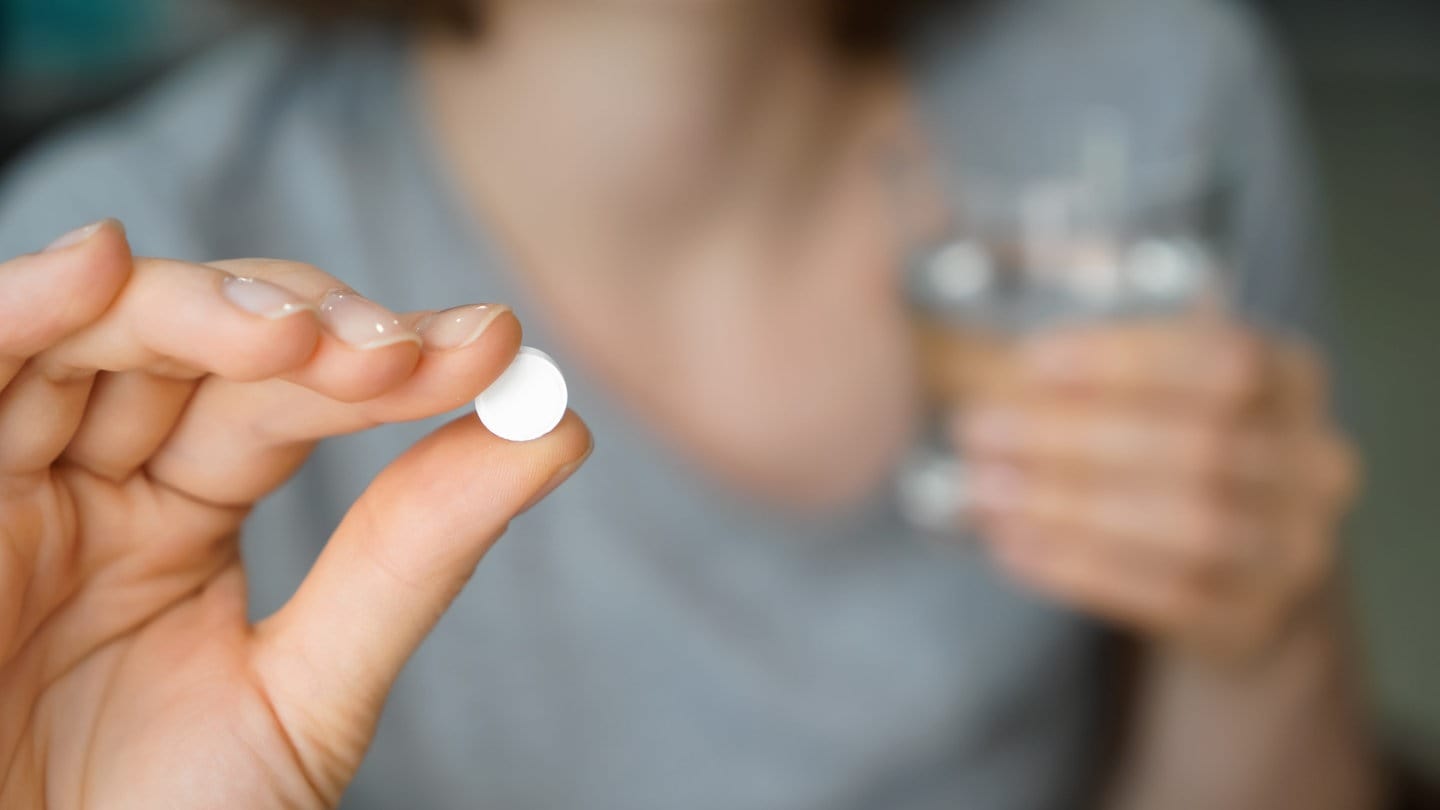 Im Vordergrund ist die Hand einer Person zu sehen, die eine weiße Pille hält. Unscharf im Hintergrund hält die Person in der anderen Hand ein Glas Wasser.