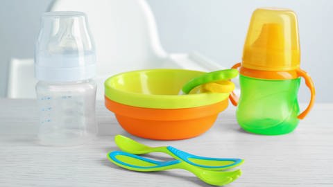 Auf einem Tisch steht buntes Plastik-Geschirr für Kinder: Mehrere Schüsseln, Löffel und zwei Trinkflaschen. Im Hintergrund ist ein Stuhl für Baby zu erkennen.