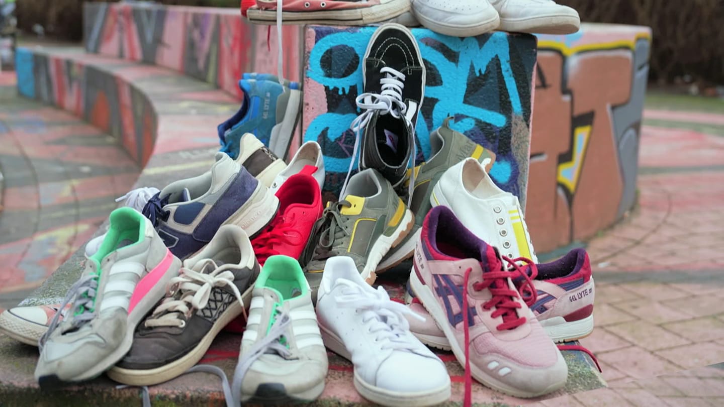 Mehrere Sneaker von Marken wie Vans, Adidas, Nike und Veja liegen auf einem Haufen auf einer mit Graffiti besprayten Mauer.