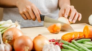Gute und scharfe Messer sind ein Muss in jeder Küche. Woran erkennt man langlebige Kochmesser? Und ist Preis gleich Qualität? Eine Frau schneidet eine Zwiebel auf einem Küchenbrett.