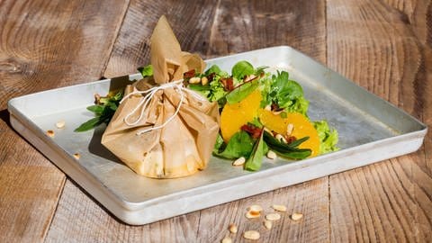 Ein zugeschnürtes Pergamentpäckchen steht neben Salat auf einem Brett